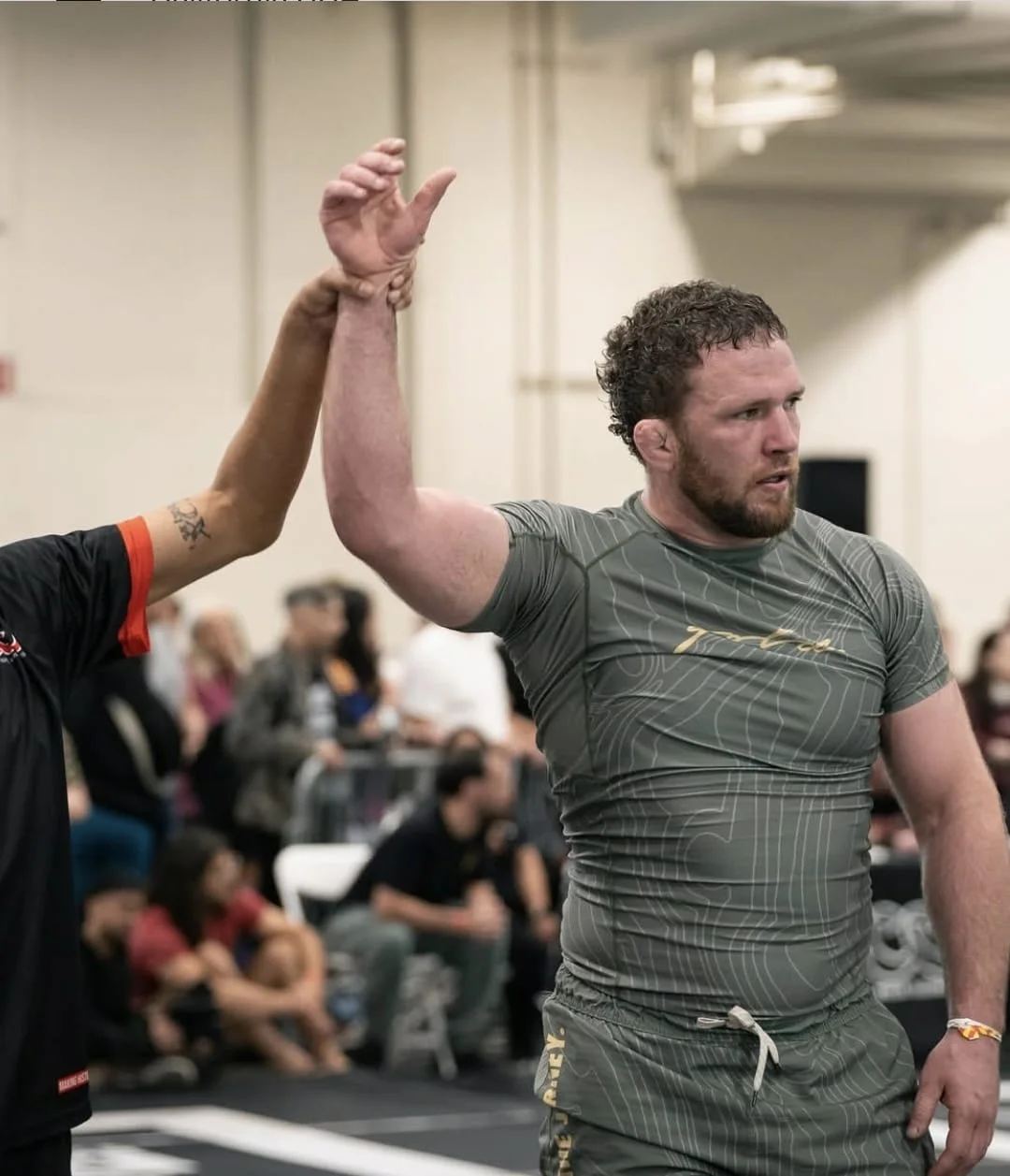 A referee raising a grappler's arm in a green topographic-print rash guard inside a competition hall with spectators behind.