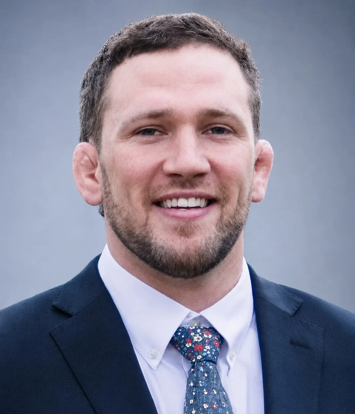 Portrait of Evan Rosborough smiling, in a navy suit, white shirt and a floral tie, against a soft gray backdrop.