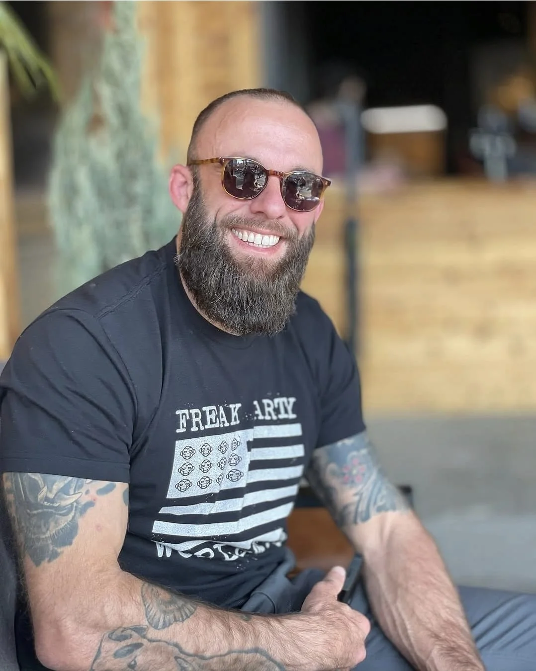 Julian smiling in tinted sunglasses and a black graphic T-shirt, seated outdoors in front of a blurred wooden storefront.