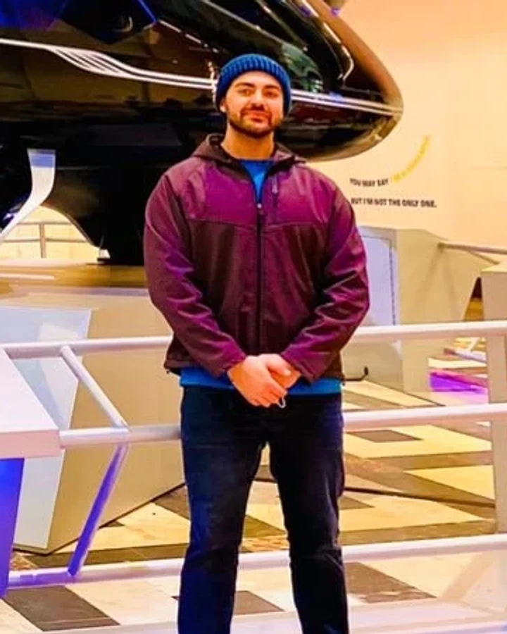 A smiling man in a plum jacket and navy beanie stands with his hands clasped in front of a glossy black air vehicle display.