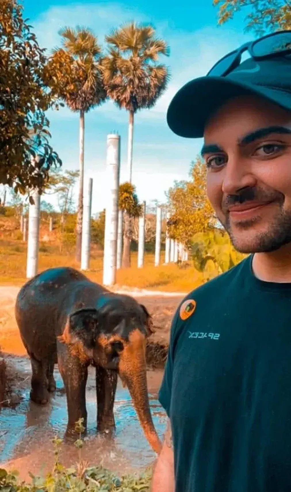 A smiling man in a dark teal cap and shirt takes a selfie with a young elephant standing in a muddy pool behind him, among tall palm trees.