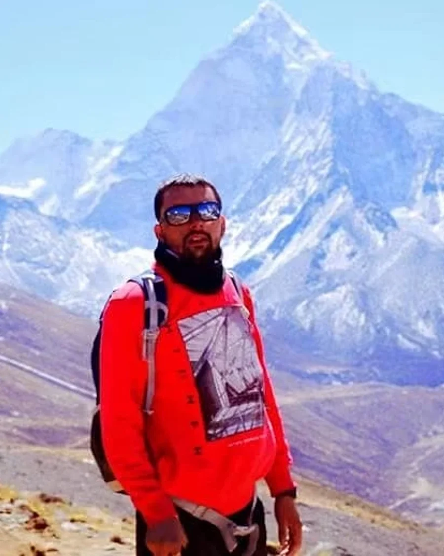 Vikas in a red jacket, sunglasses and a backpack, standing in front of a snow-capped mountain peak.