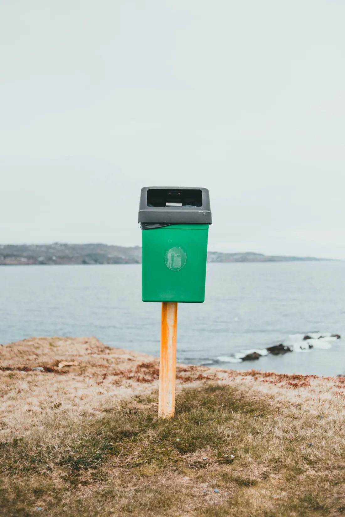A green waste bin on a post on a grassy headland above a gray bay.