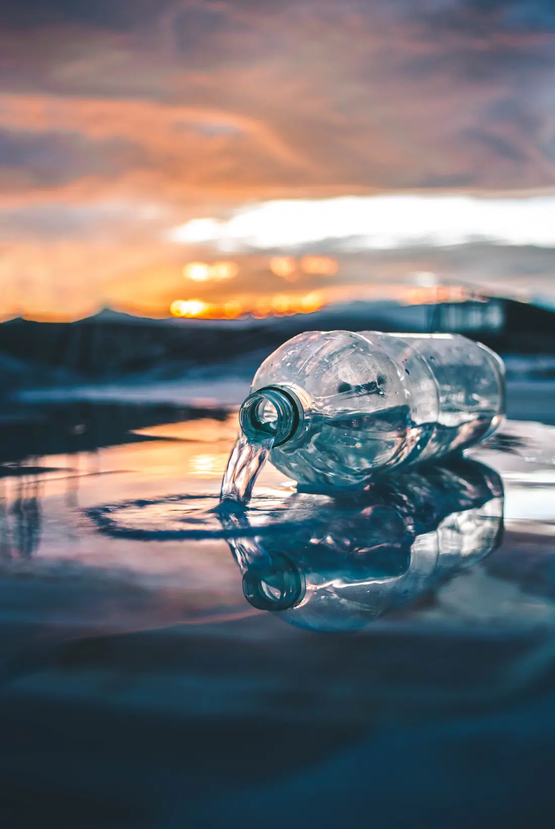 A clear plastic bottle lying in shallow water at sunset, with its reflection below.