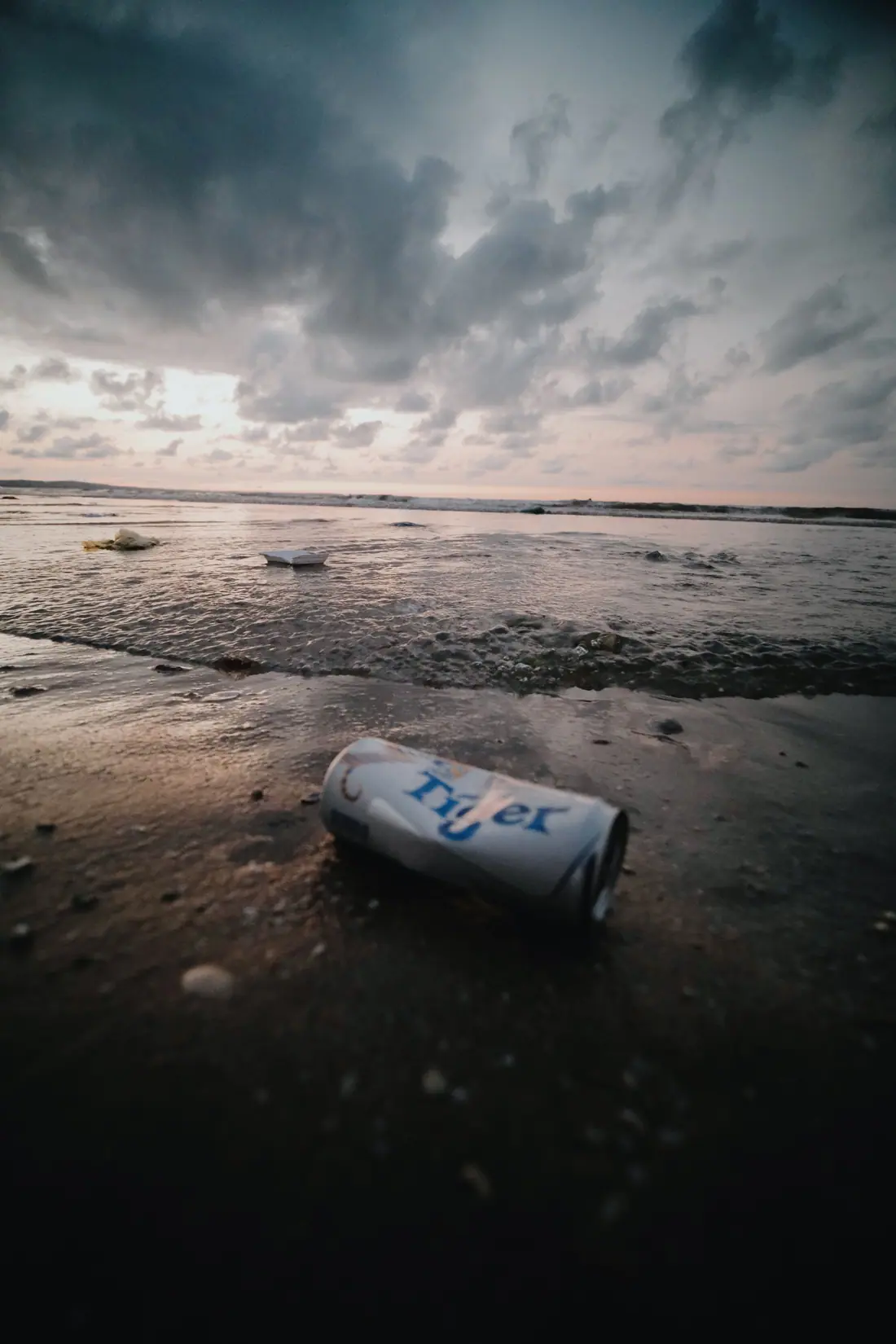 A crushed can lying on wet sand at dusk beneath heavy clouds, with litter floating in the surf.