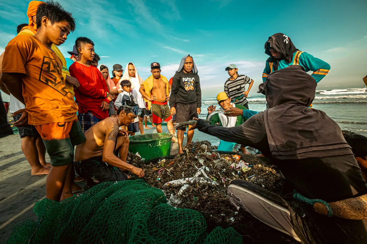 A crowd of people on a beach gathered around a net of mixed waste and fish as two people sort through it.