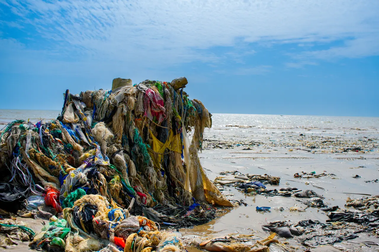 A tall heap of fabric, netting and plastic waste on a tidal beach under a blue sky.