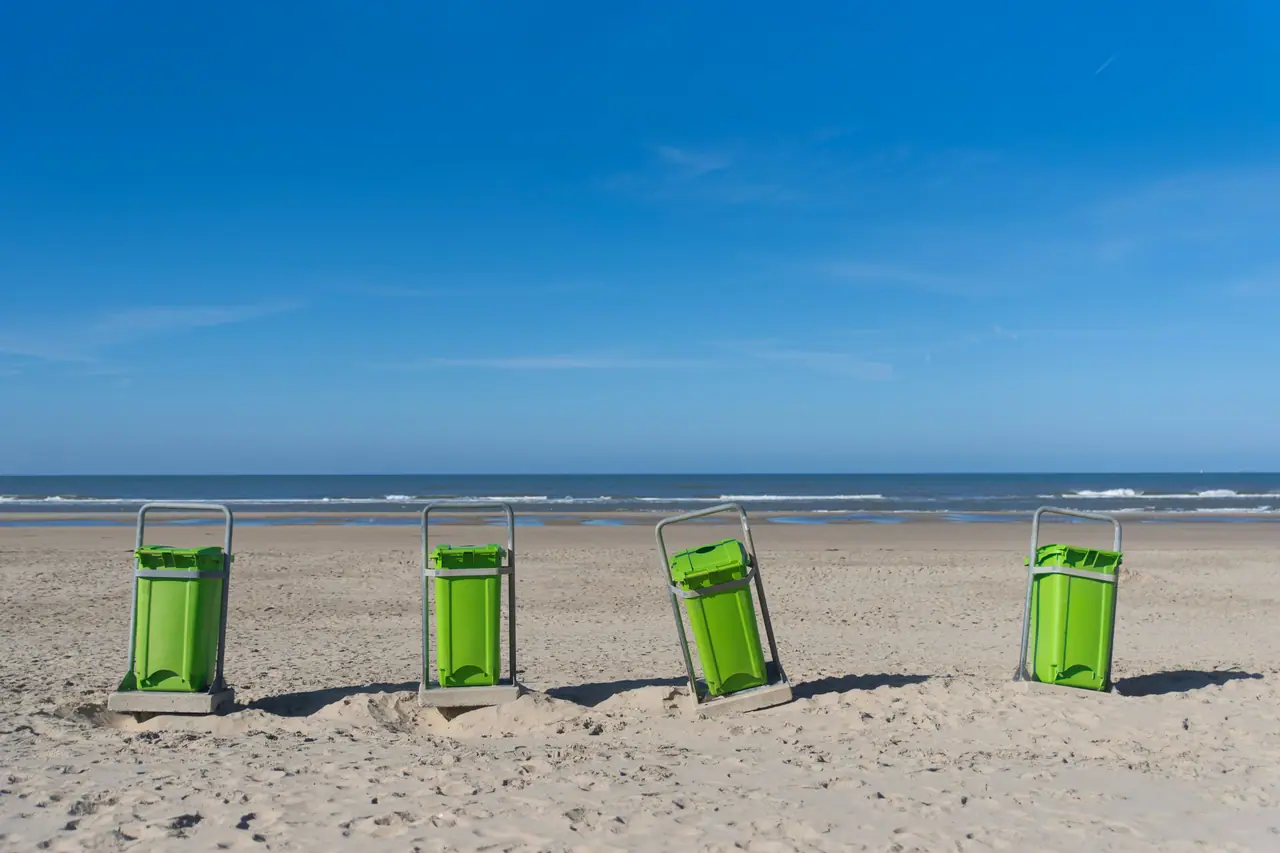 Four bright green waste bins standing in a row on an open beach beneath a deep blue sky.