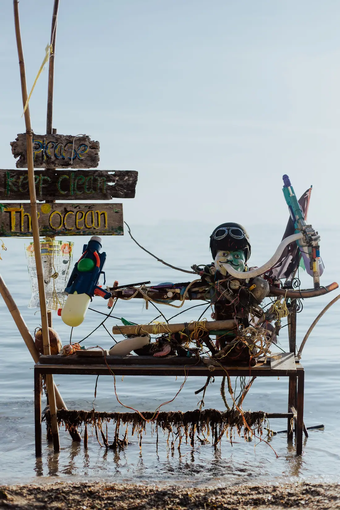 A hand-painted driftwood sign reading Please keep clean the ocean, beside an assemblage of washed-up objects standing in shallow water.