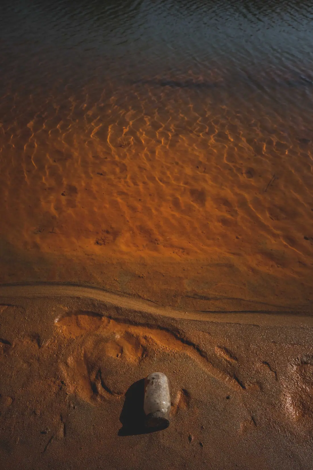 A glass jar lying on the sand beside shallow, copper-coloured water with rippling patterns.