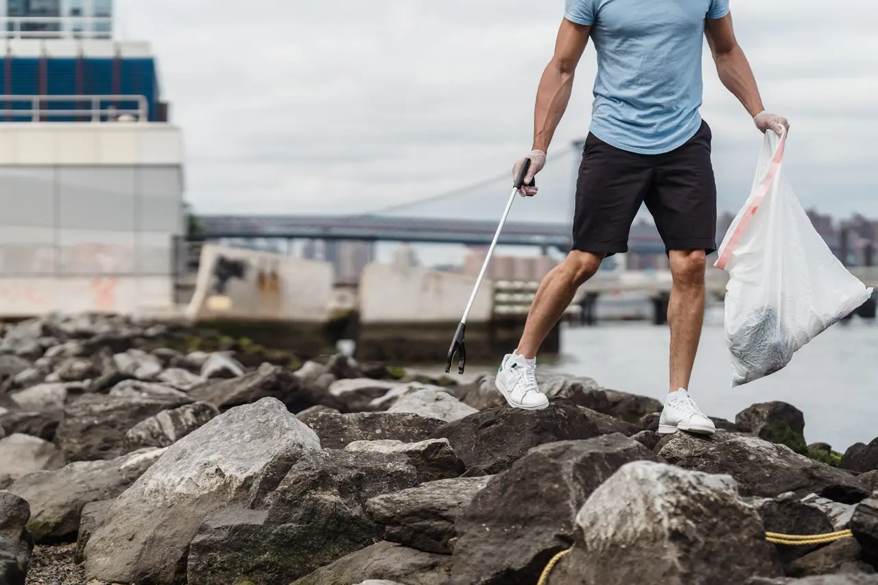 A volunteer in gloves holding a litter grabber and a bag, standing on rocks beside a waterfront with a bridge in the distance.